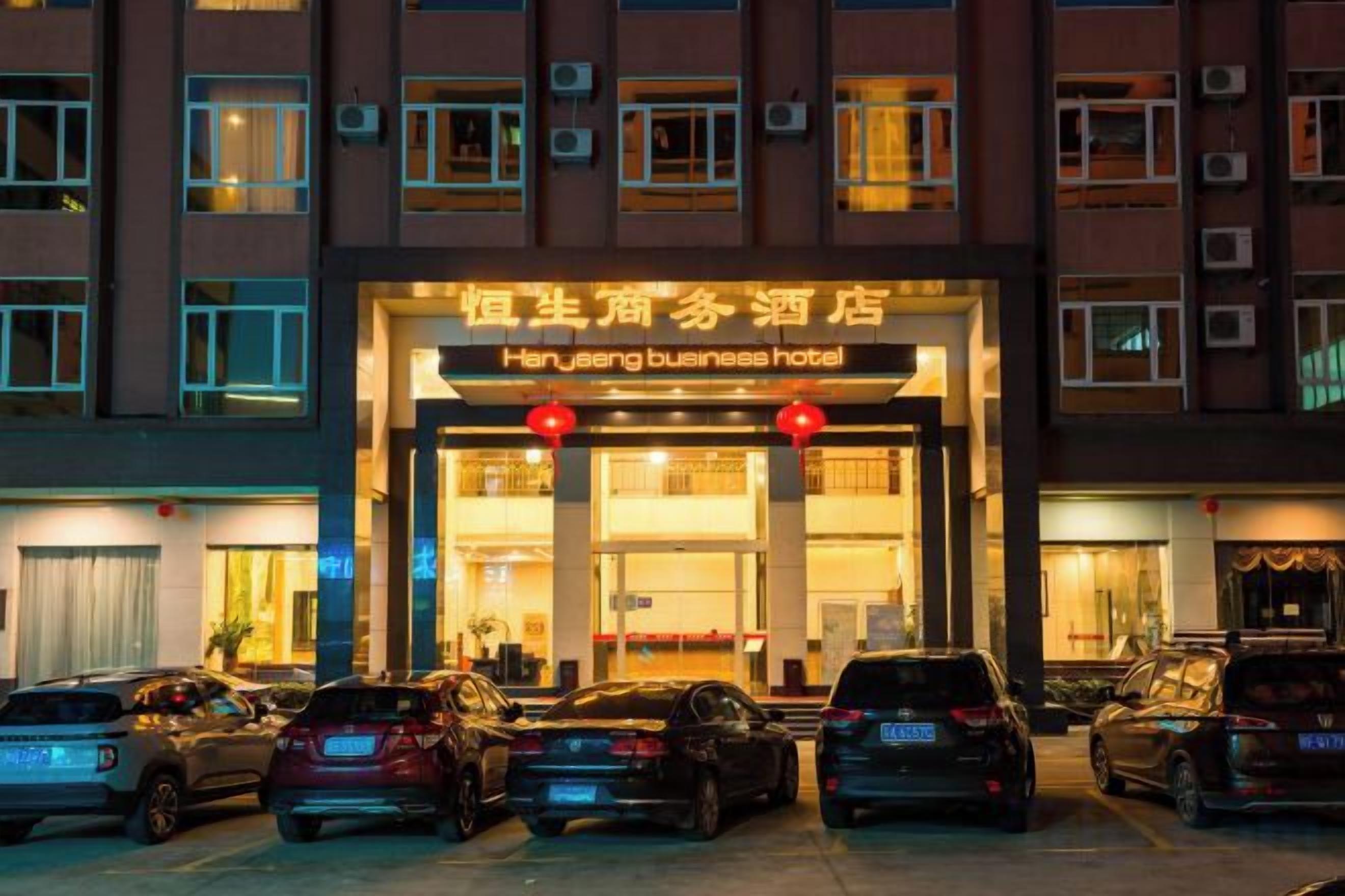 A hotel entrance with a sign, multiple parked cars, and a building facade with windows and air conditioning units.