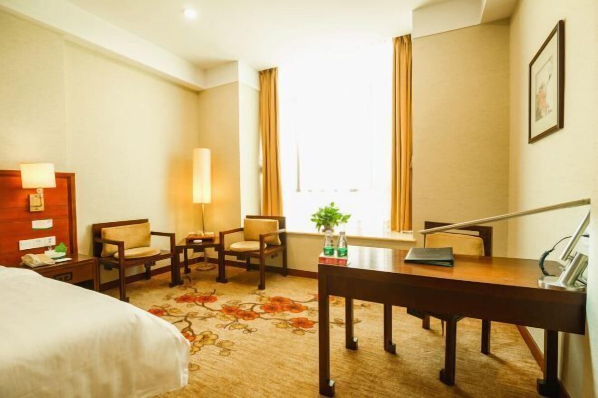 Una habitación de hotel con cama, escritorio, dos sillas, lámpara, ventana con cortinas y alfombra estampada.