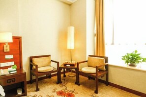 Habitación de hotel con escritorio de madera, dos sillas, una mesita, lámpara, ventana con cortinas y una planta en el alféizar.