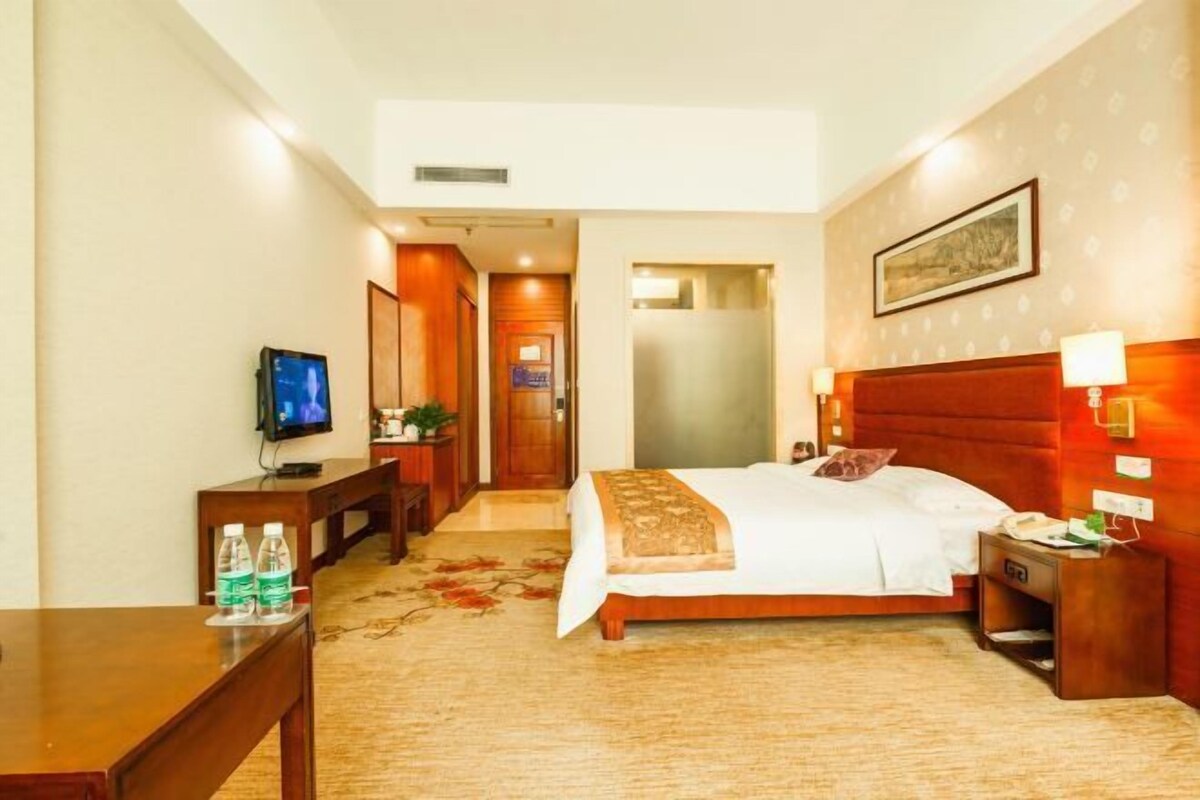 Una habitación de hotel con cama, mesita de noche, lámpara y televisión sobre un escritorio.
