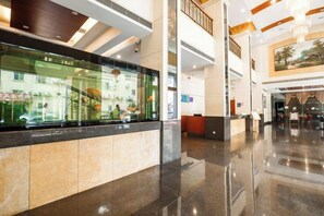 Un vestíbulo de hotel con una recepción, una vitrina de vidrio y una escalera que sube a un nivel superior.