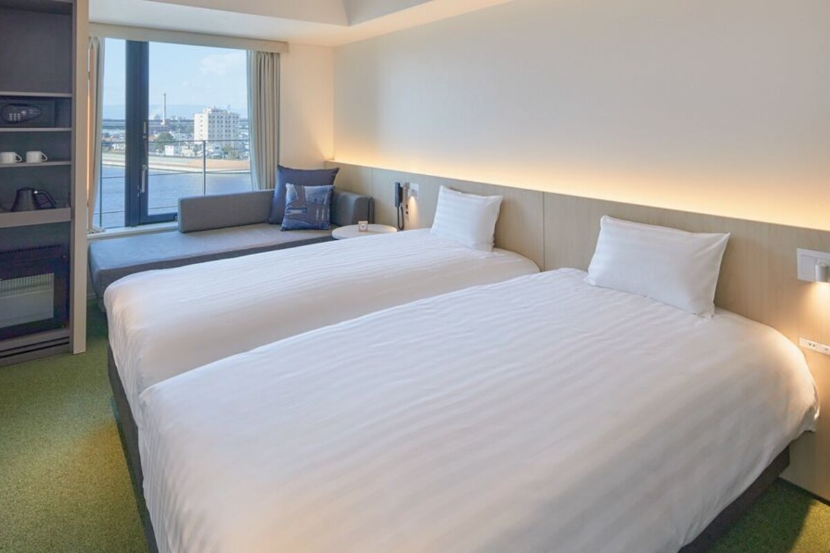Habitación de hotel con dos camas, zona de estar con vistas y estante empotrado con dispositivos electrónicos.