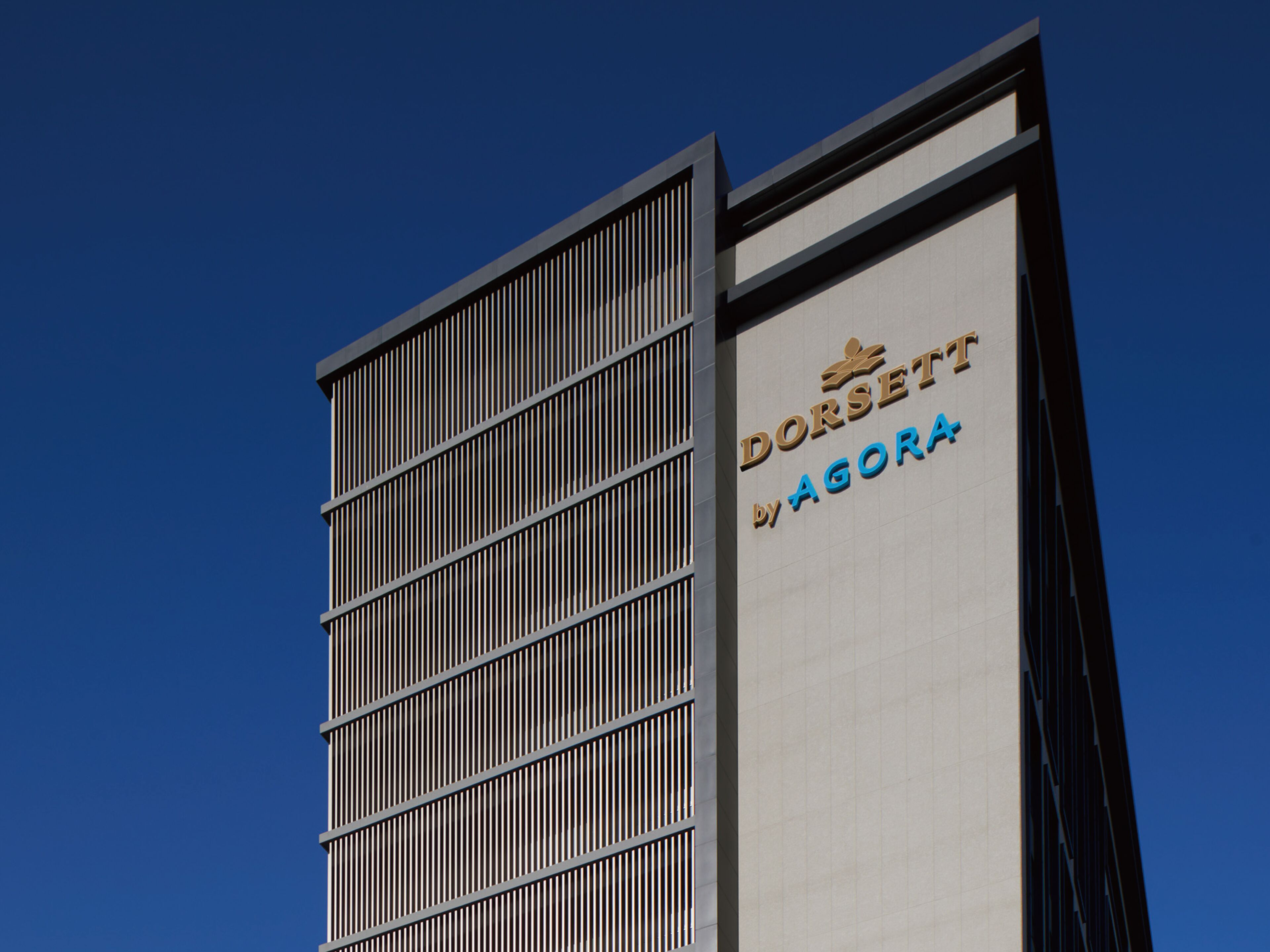 A modern building with a vertical facade featuring a series of vertical slats. The name "DORSETT by AGORA" is prominently displayed on the side.