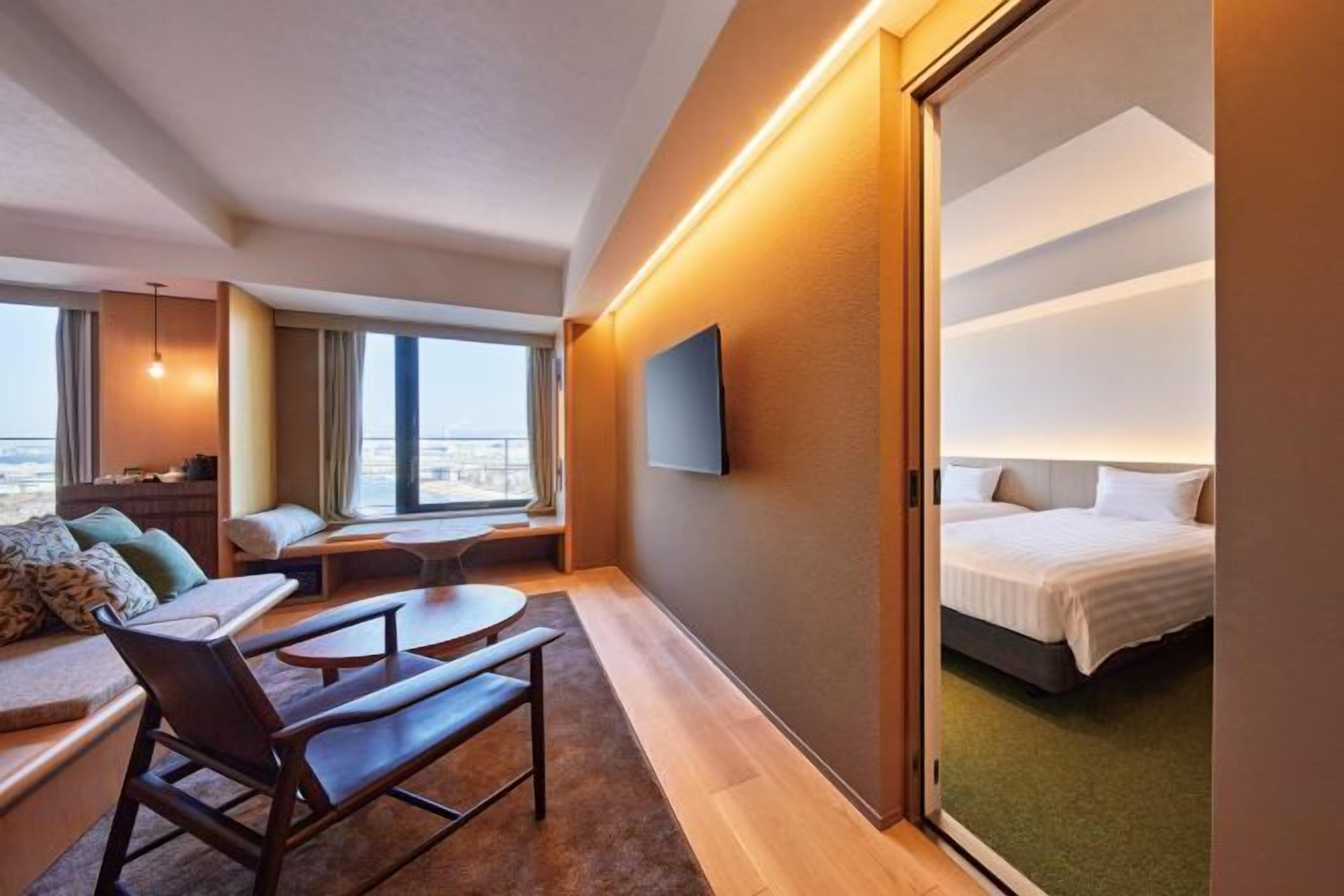 A modern hotel room with a large bed, a seating area with a table, a TV mounted on the wall, and a view of the city through the window.