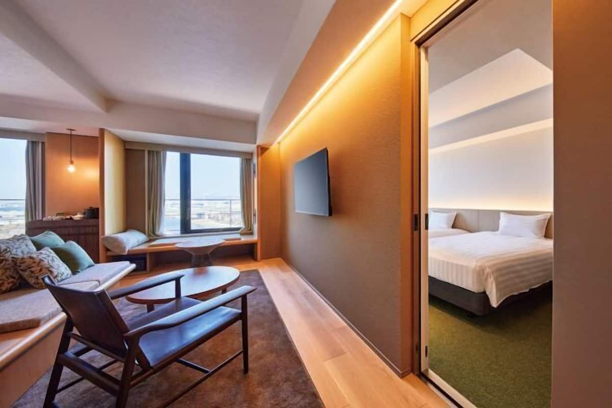 A modern hotel room with a large bed, a seating area with a table, a TV mounted on the wall, and a view of the city through the window.