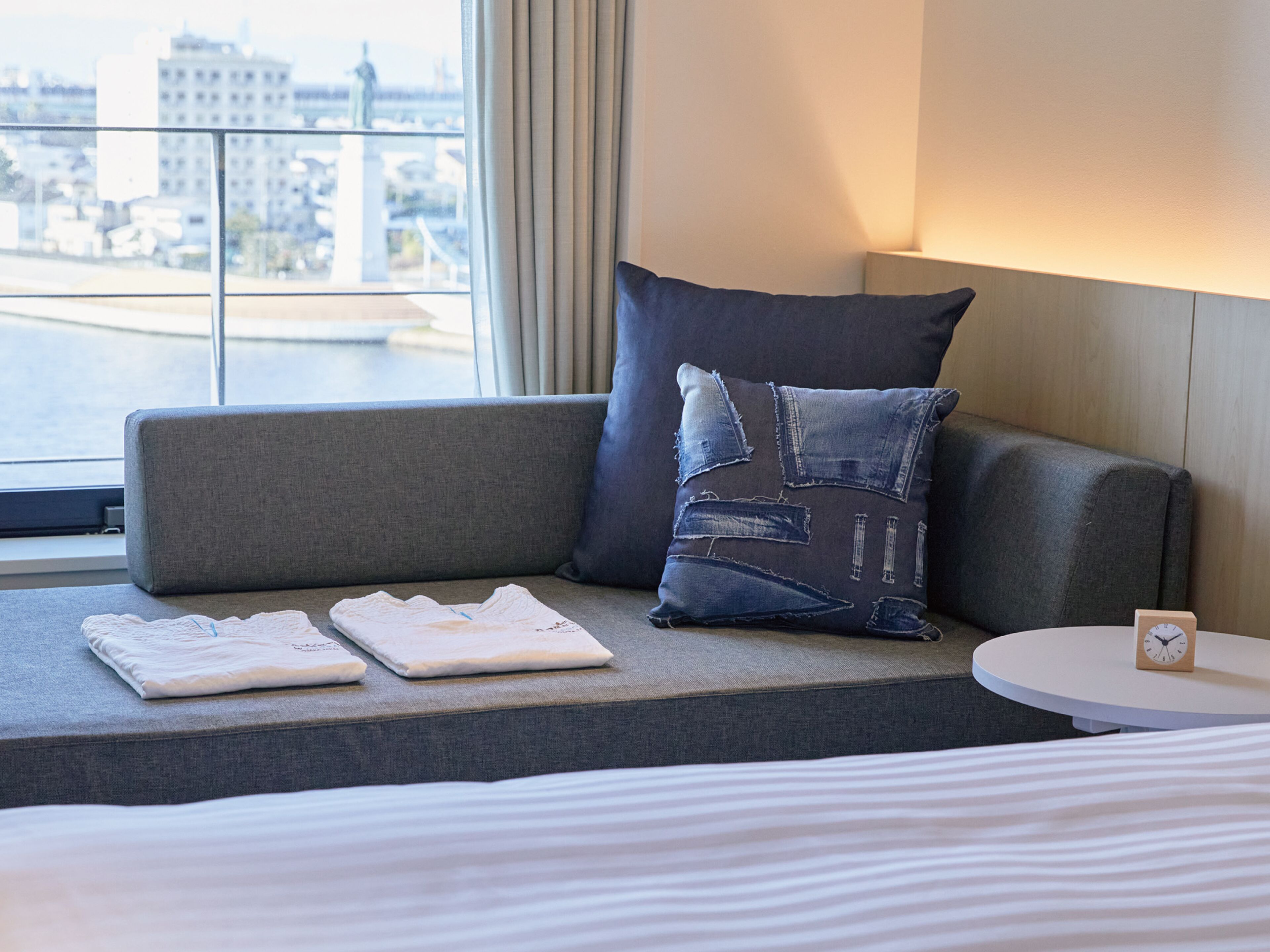 Habitación de hotel con un sofá gris, un cojín con diseño vaquero, una mesita redonda con un reloj y vistas a un paisaje urbano a través de una ventana.