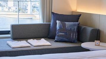 Habitación de hotel con un sofá gris, un cojín con diseño vaquero, una mesita redonda con un reloj y vistas a un paisaje urbano a través de una ventana.