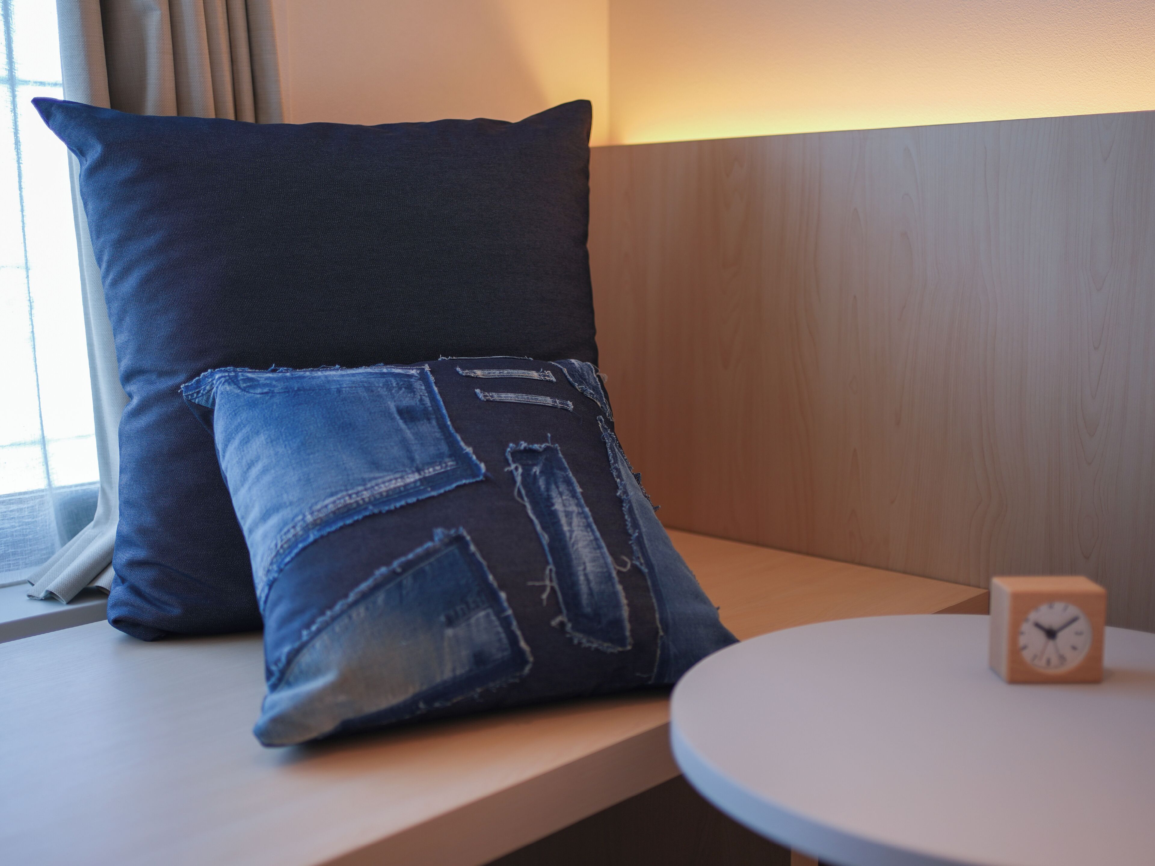 Una cama con un cojín azul oscuro y un cojín más pequeño con parches de mezclilla, un cabecero de madera y una mesa redonda con un reloj.