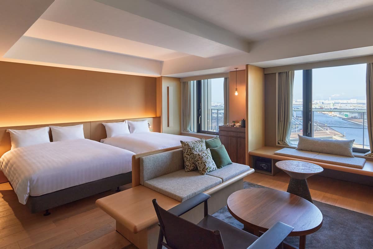 Habitación de hotel con dos camas, un sofá, una mesita y vistas a la ciudad a través de amplias ventanas.