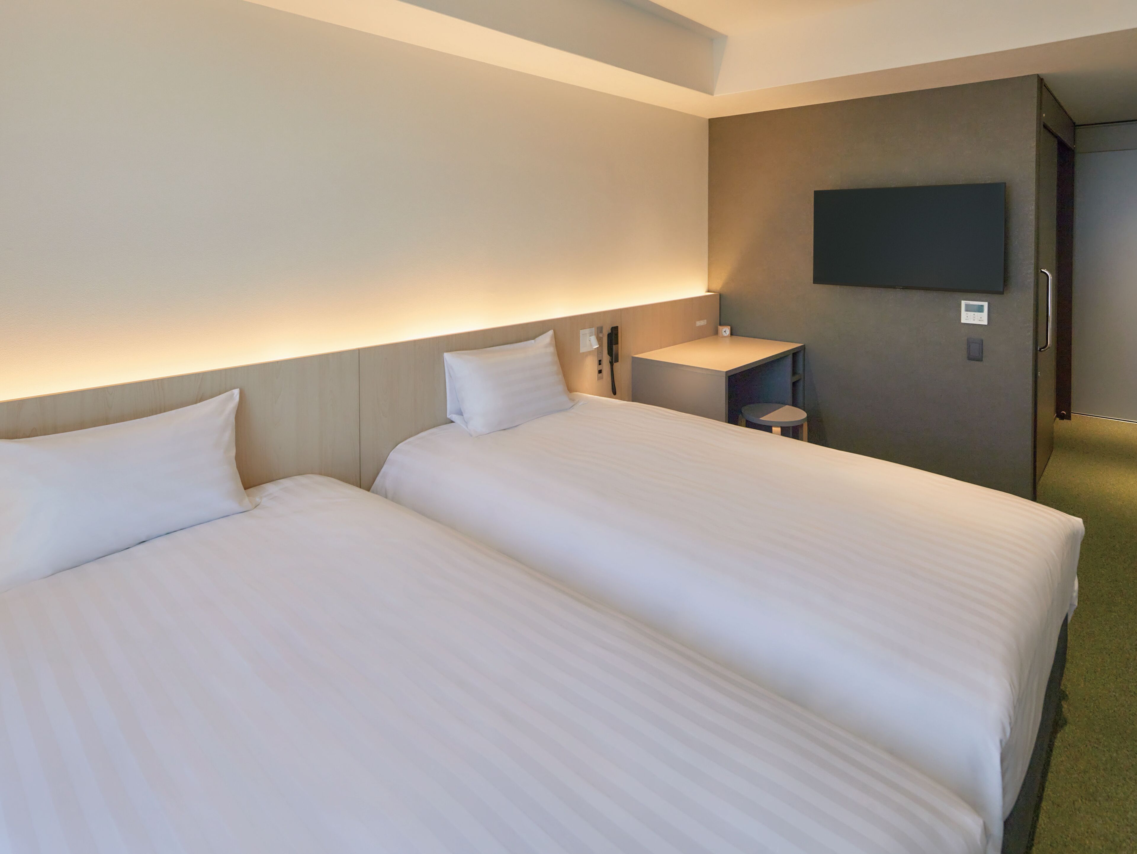 A hotel room with two beds, a bedside table, a small stool, a flat-screen TV mounted on the wall, and a thermostat.