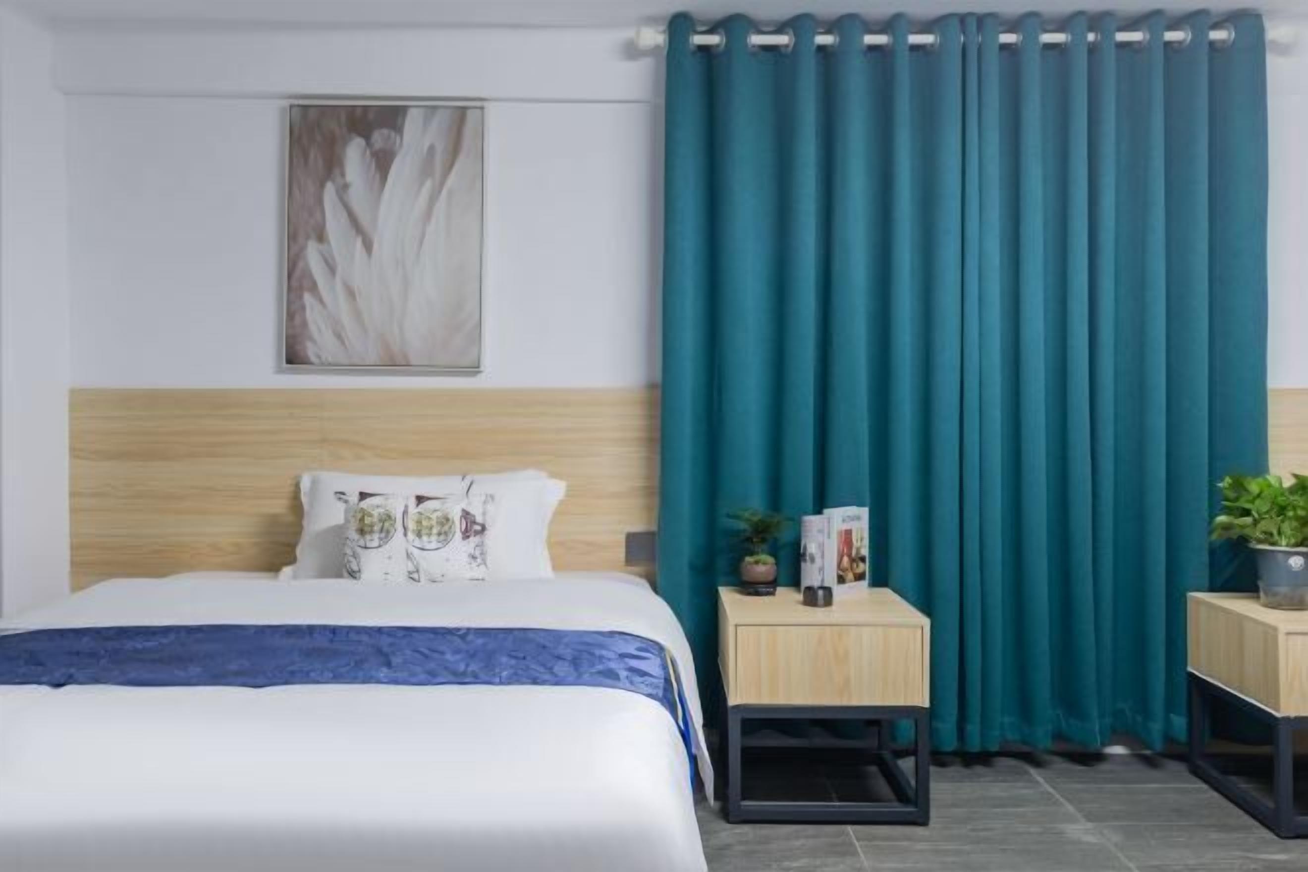 A hotel room with a bed, a headboard, a nightstand with a plant, a framed picture on the wall, and teal curtains.