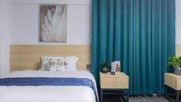 A hotel room with a bed, a headboard, a nightstand with a plant, a framed picture on the wall, and teal curtains.