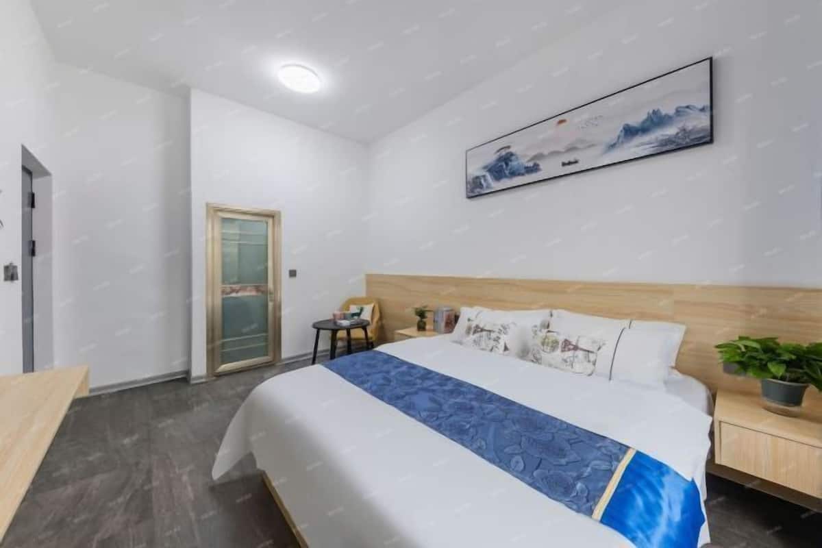 A hotel room with a bed, a nightstand, a chair, a small table, a potted plant, and a painting on the wall.