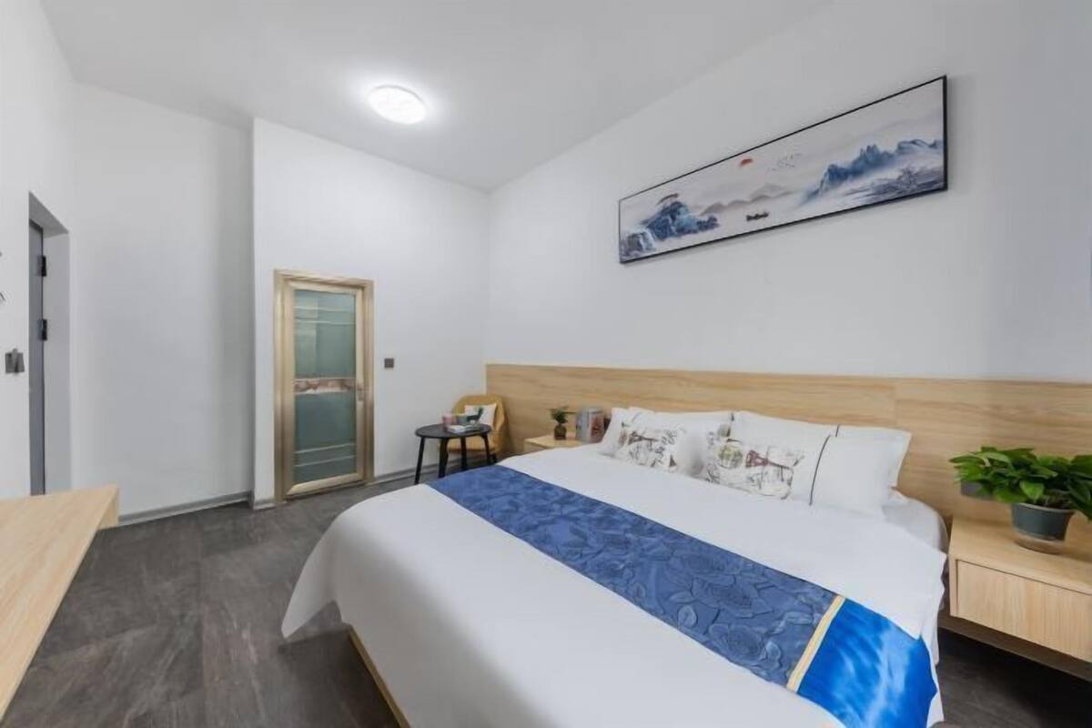 A hotel room with a bed, a nightstand, a chair, a small table, a potted plant, and a painting on the wall.