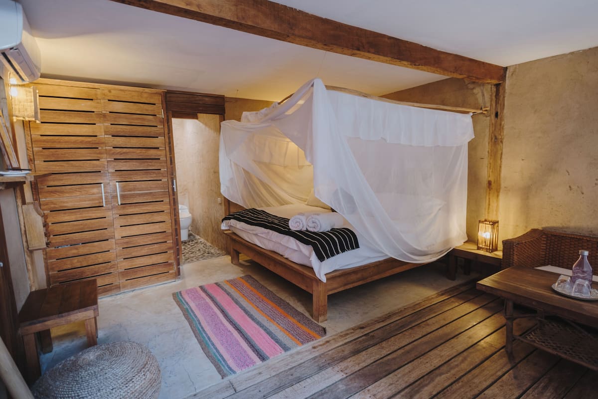 A wooden bed with a canopy, a small table, a wicker chair, and a colorful striped rug.