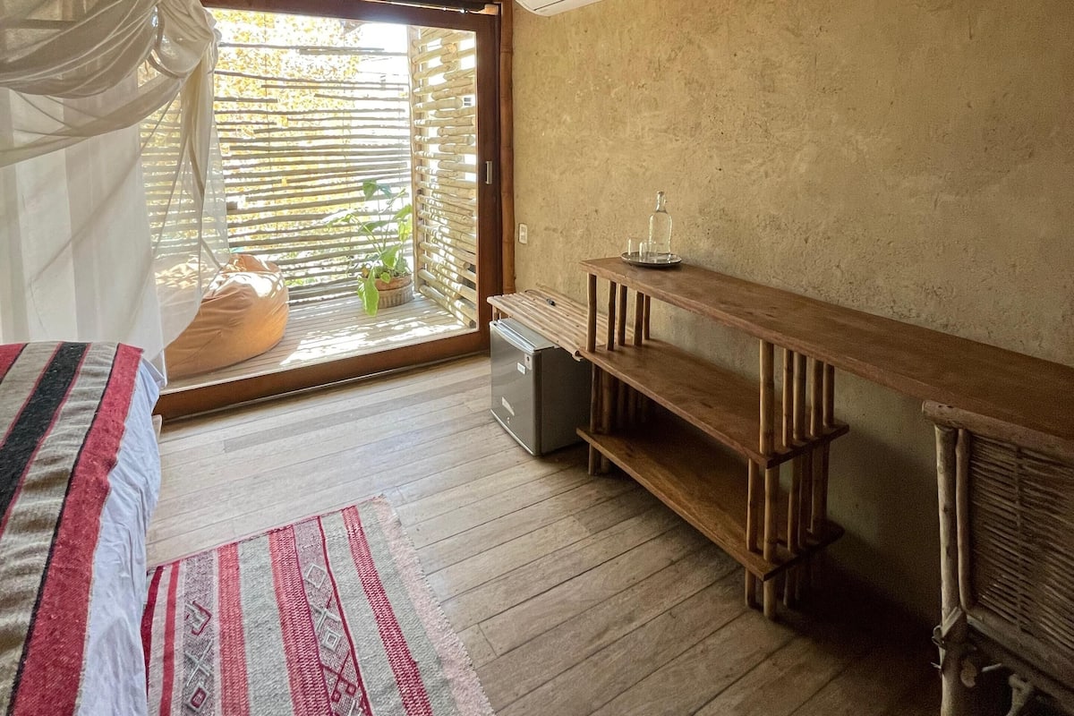 A room with a bed, a wooden desk, a small refrigerator, and a window with a view of greenery.