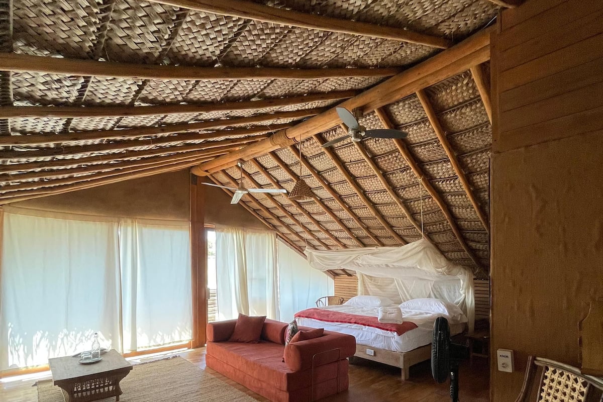 A room with a high wooden ceiling, a bed with white bedding, a red sofa, a small table, and a chair.