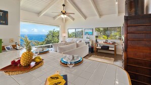 A spacious living room with a large window overlooking the ocean, a ceiling fan, and a wooden door. - Tranquil Ocean View Cottage: Your Idyllic Escape at Waipio Valley, Big Island HI (Honokaa)