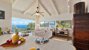 A spacious living room with a large window overlooking the ocean, a ceiling fan, and a wooden door.