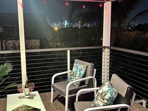 A balcony with two white metal chairs, a table with two wine glasses and a bottle, and a view of a cityscape at night.