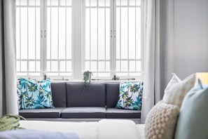 Un moderno salón con un sofá gris, dos ventanas con marcos blancos y vistas al mar.