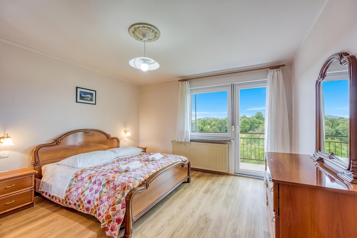 Quarto com cama de madeira, mesa de cabeceira, cômoda, janela com cortinas e vista para o verde.