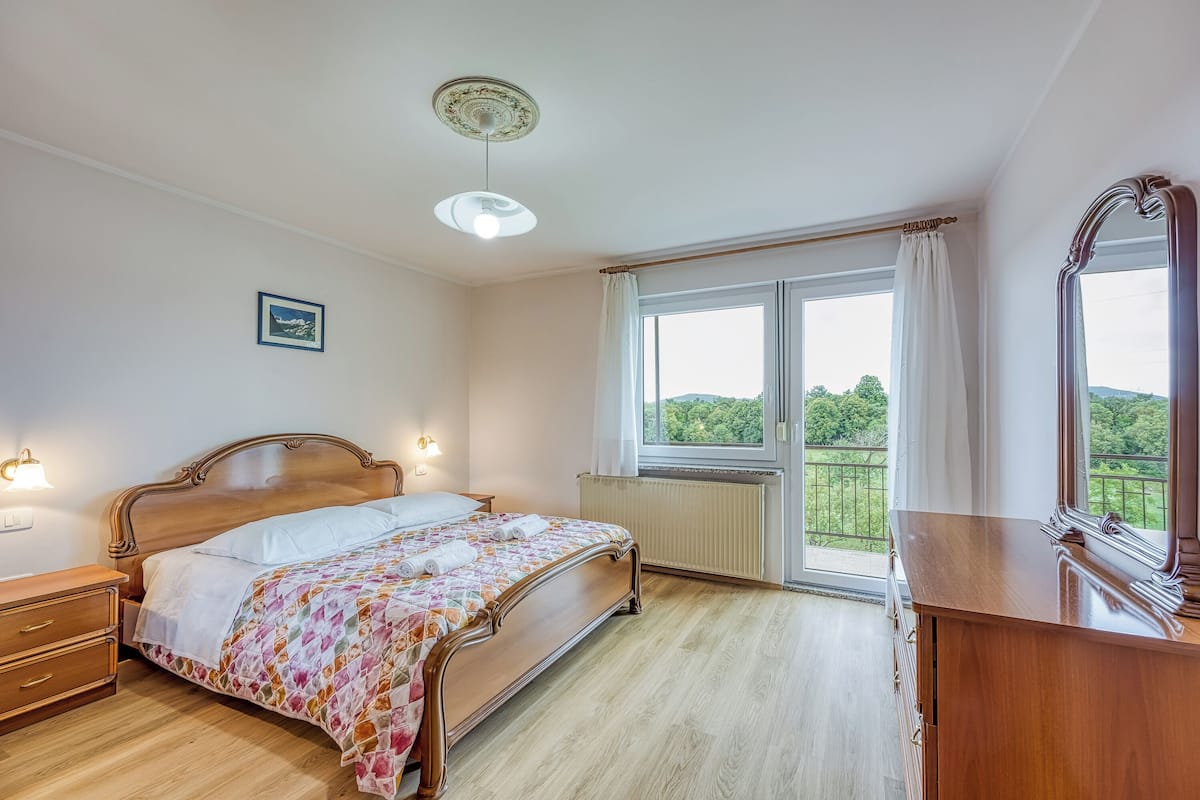 Une chambre à coucher avec un lit en bois, une table de chevet, une commode, une fenêtre donnant sur un paysage verdoyant et un luminaire au plafond.