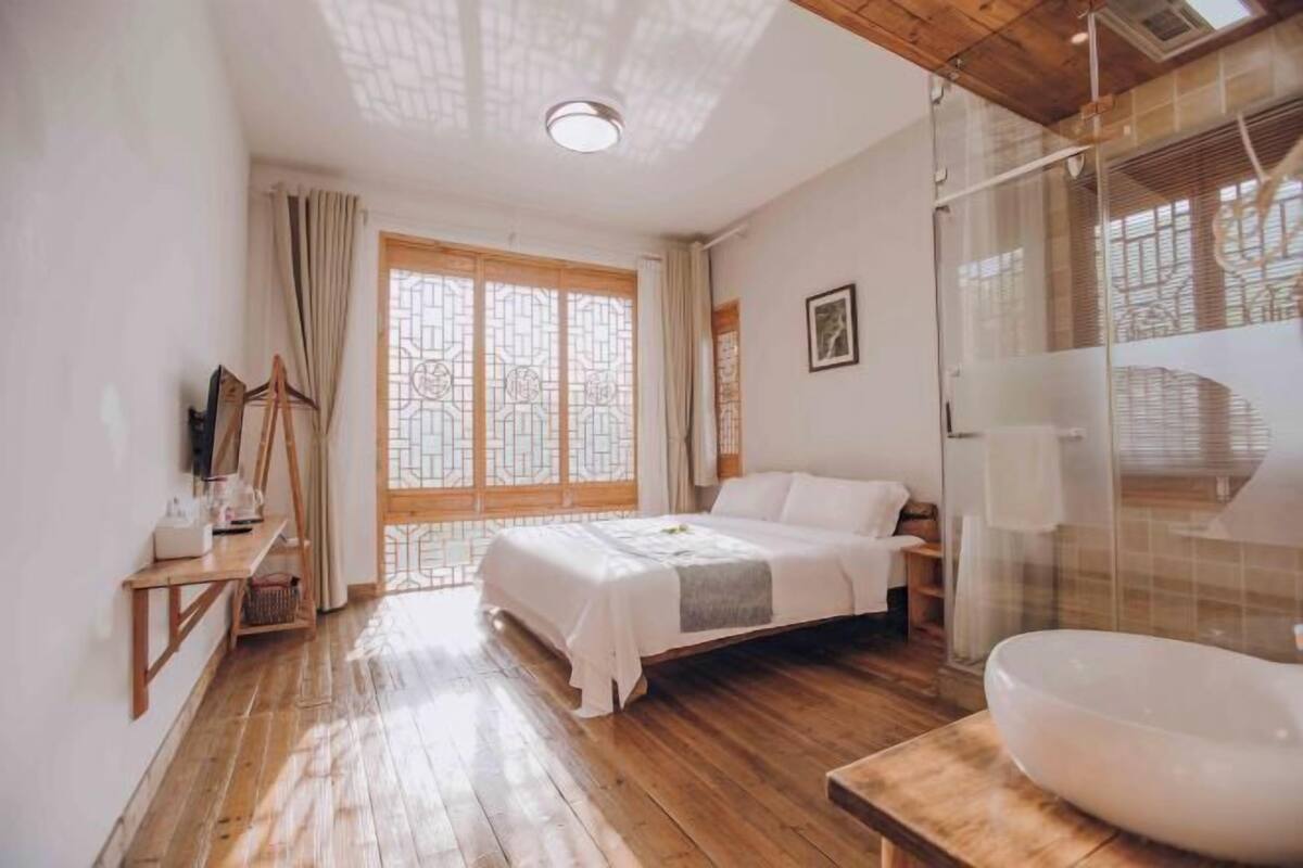 A bedroom with a wooden floor, a bed with white linens, a bedside table, a large window with a view, and a bathroom with a large sink.