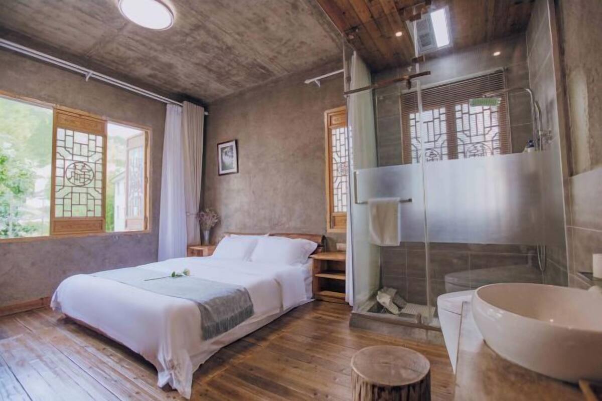 A bedroom with a large bed, a wooden floor, a glass shower enclosure, and a large white basin.