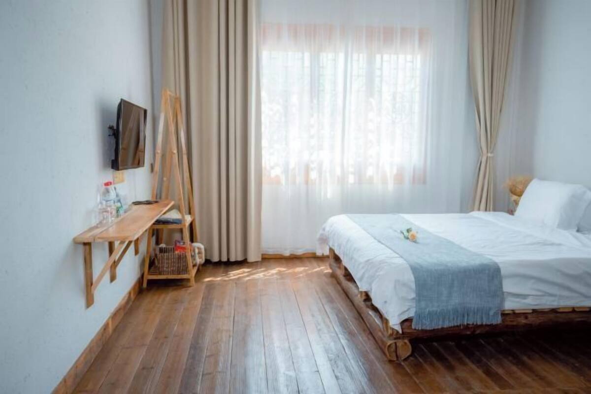 A room with a wooden bed, a small desk, a television mounted on the wall, and a window with curtains.