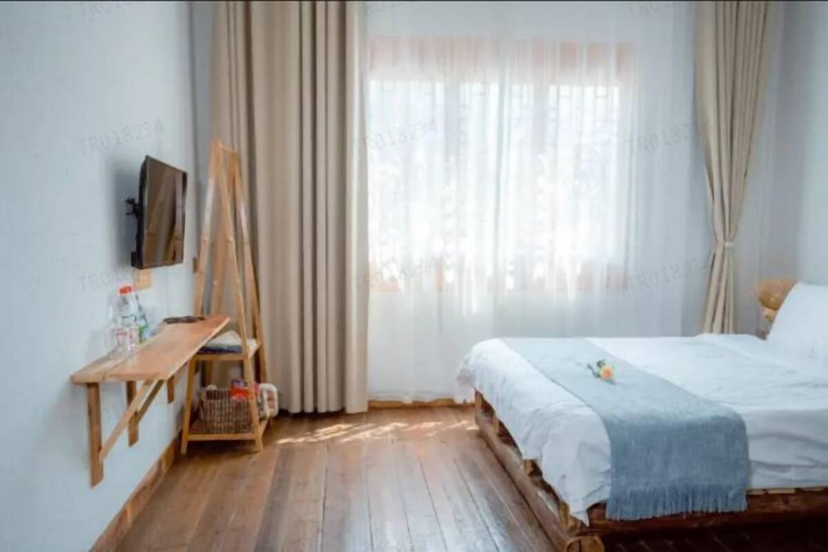 A bedroom with a wooden bed, a small wooden desk, a TV mounted on the wall, and a window with sheer curtains.