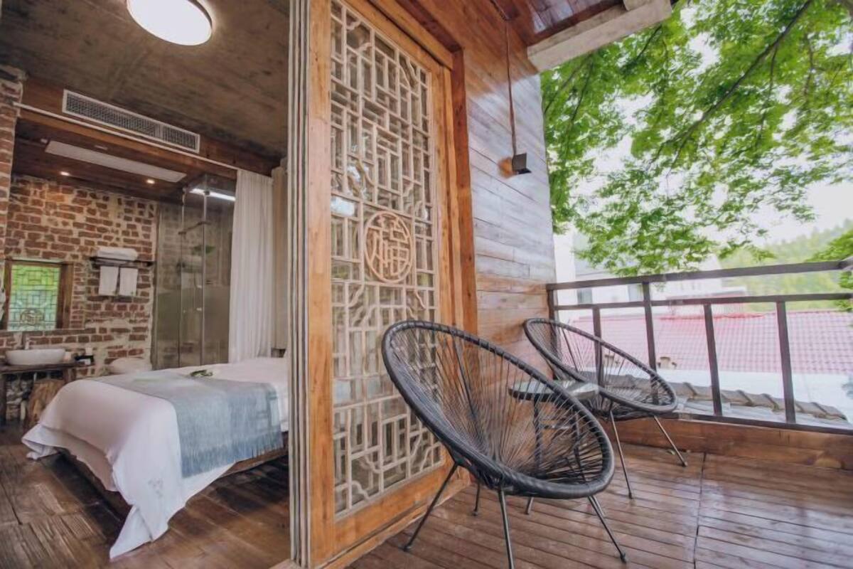 A room with a bed, a wooden chair, a decorative screen, and a view of greenery outside.