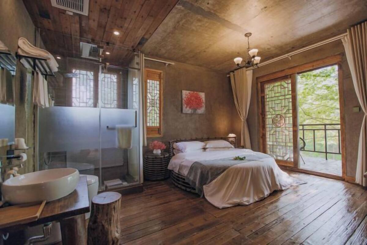 A wooden cabin bedroom with a large bed, a bathroom with a sink and shower, and a view of greenery through the window.