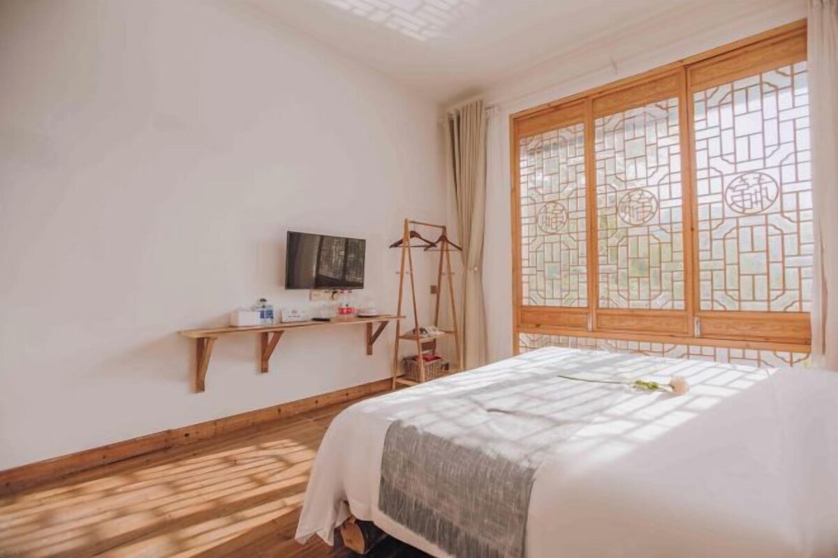A room with a bed, a TV on a shelf, a wooden desk, and a window with a decorative screen.