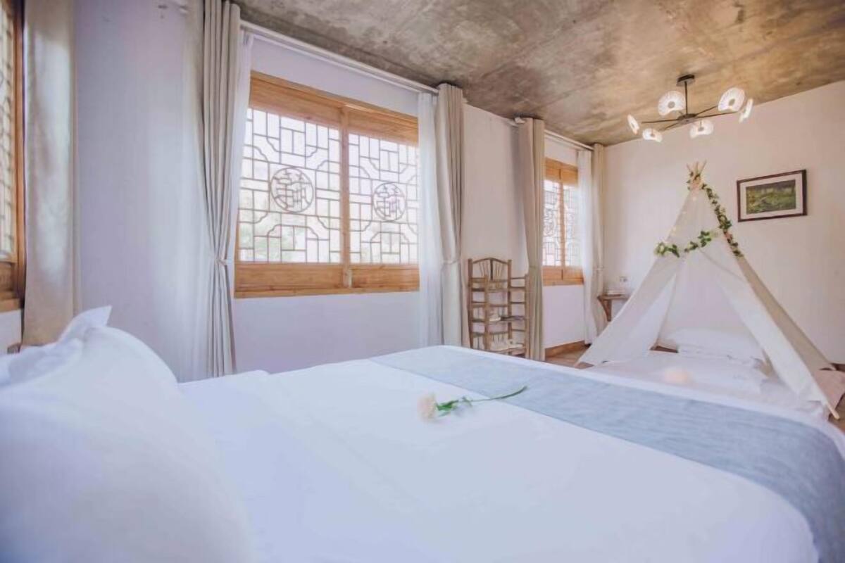 A bedroom with a bed, curtains, a window with a geometric pattern, a wooden chair, and a tent-like structure.