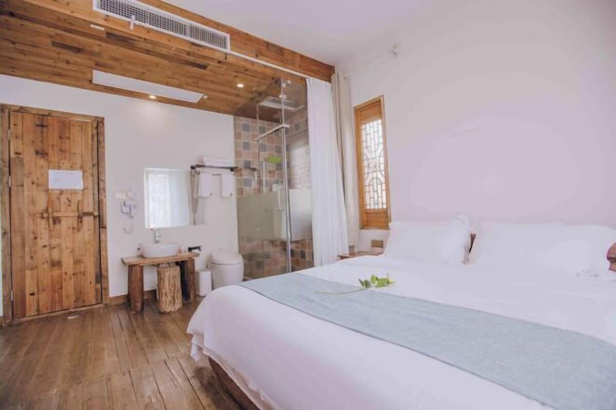 A bedroom with a wooden ceiling, a large bed, a bedside table, a mirror, and a window with curtains.