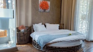 Un letto rotondo con materasso e cuscini bianchi, circondato da una struttura in legno. Al muro, un quadro raffigura un albero, e a destra si trova una finestra con tende.