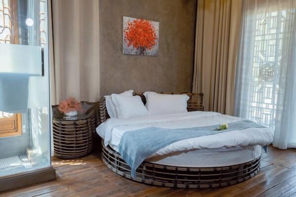A circular bed with a white mattress and pillows, surrounded by a wooden frame. There is a painting of a tree on the wall, and a window with curtains on the right. - Jushe village Meisu Inn (Deqing)