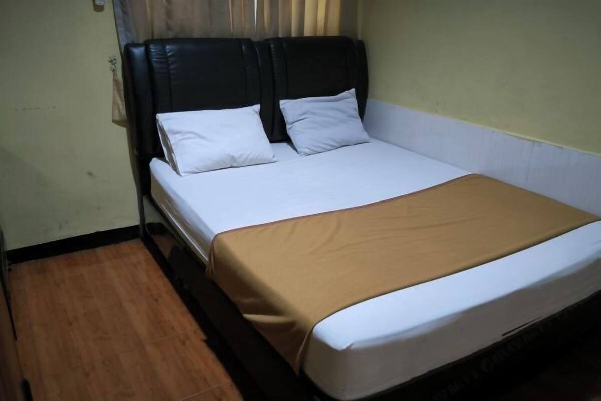 A single bed with a black headboard, white pillows, and a brown bedspread in a room with wooden flooring and light-colored walls.