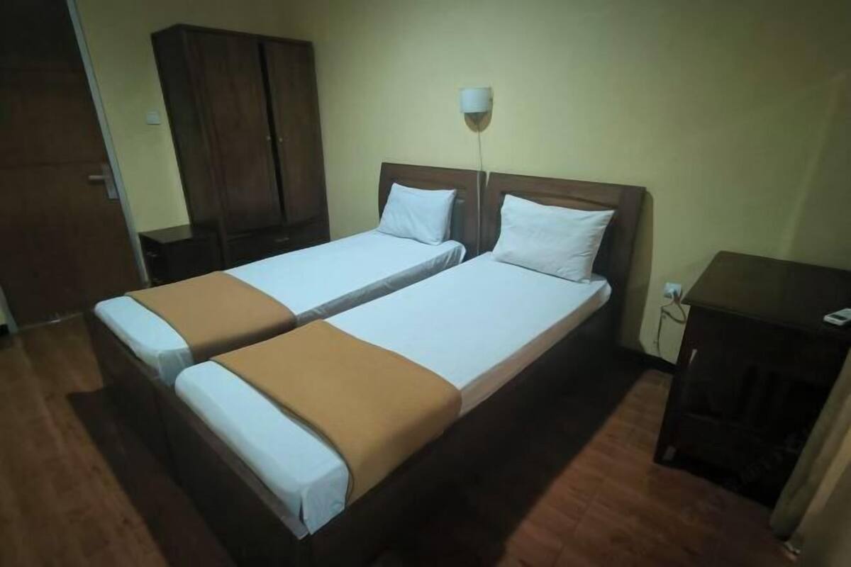 A room with two single beds, a wooden wardrobe, a bedside table, and a wall-mounted lamp.