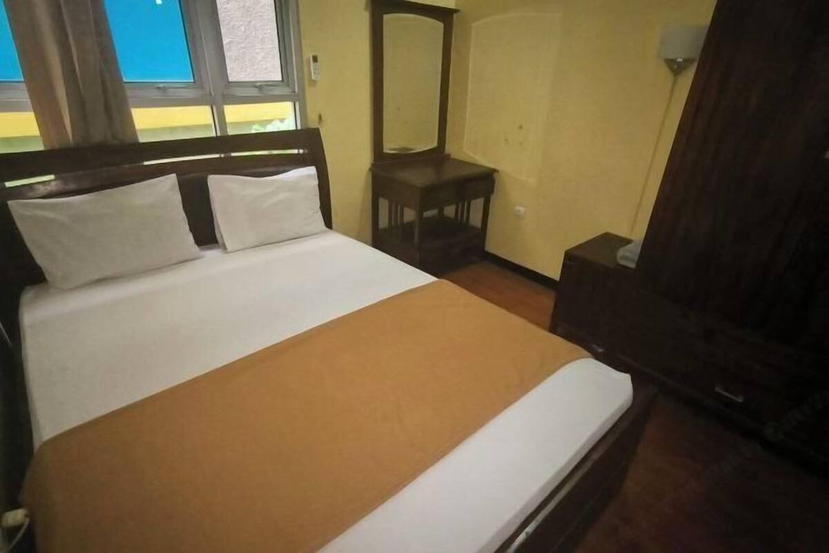 A bed with white and brown bedding, a wooden headboard, a small wooden nightstand, a mirror, and a window with a view of a building.