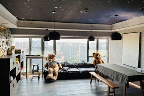 A modern living room with a black leather sofa, two teddy bears, a dining table, and a city view through large windows. - Bonjour Youth Hostel (Shenzhen)