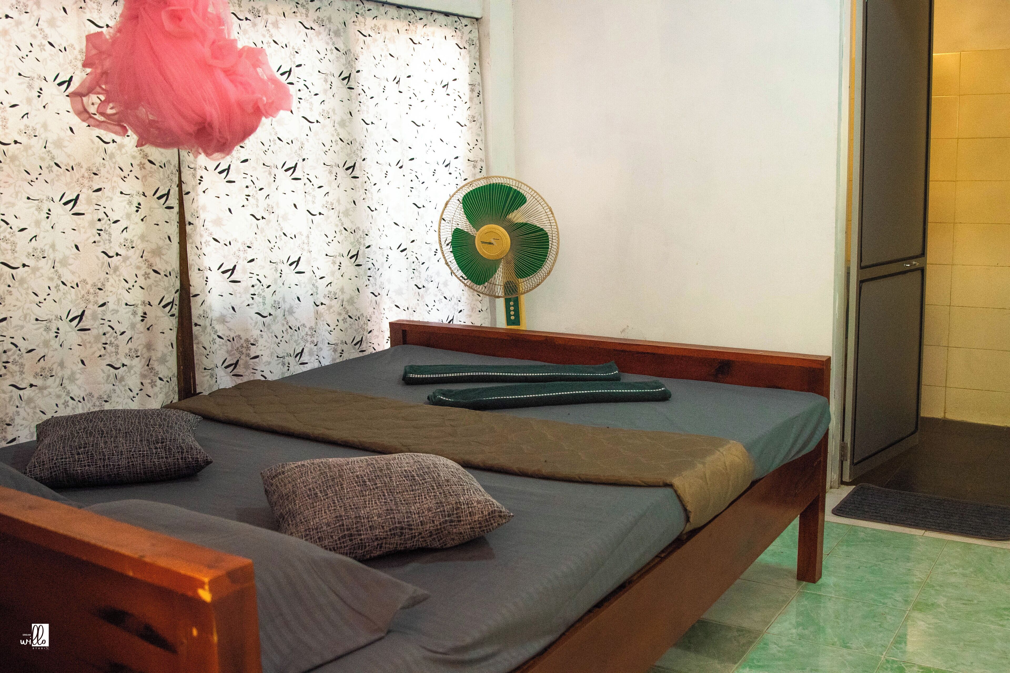 Een slaapkamer met een bed, kussens, een ventilator en een raam met gordijnen met een patroon.