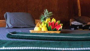 A bed with a wooden headboard, a green blanket, and a fruit basket containing pineapples, bananas, and watermelon. - Ynot Udawalawa Safari & Resort (Udawalawa)