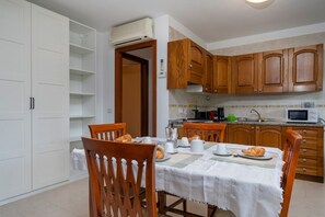 A dining area with a table set for four, wooden chairs, a white tablecloth, and a kitchen in the background with wooden cabinets and a microwave.