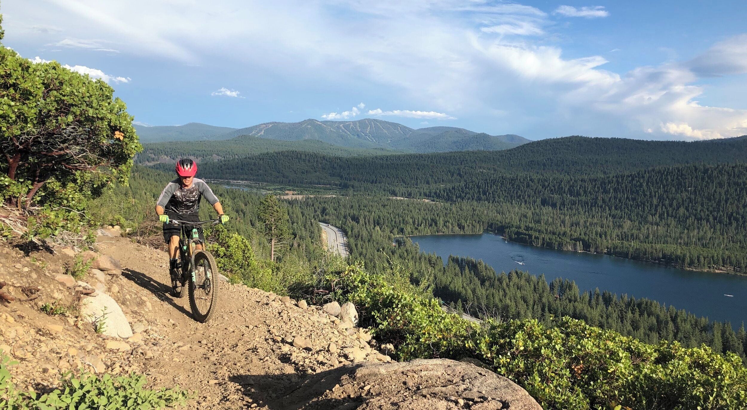 Un ciclista de montaña en un sendero de tierra con un lago y montañas boscosas al fondo.