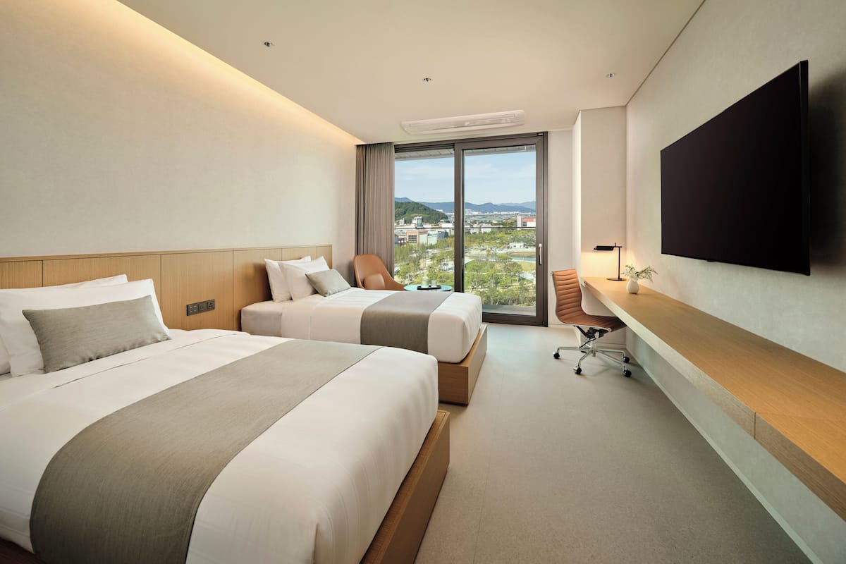 A hotel room with two beds, a desk, a chair, a flat-screen TV, and a large window offering a city view.