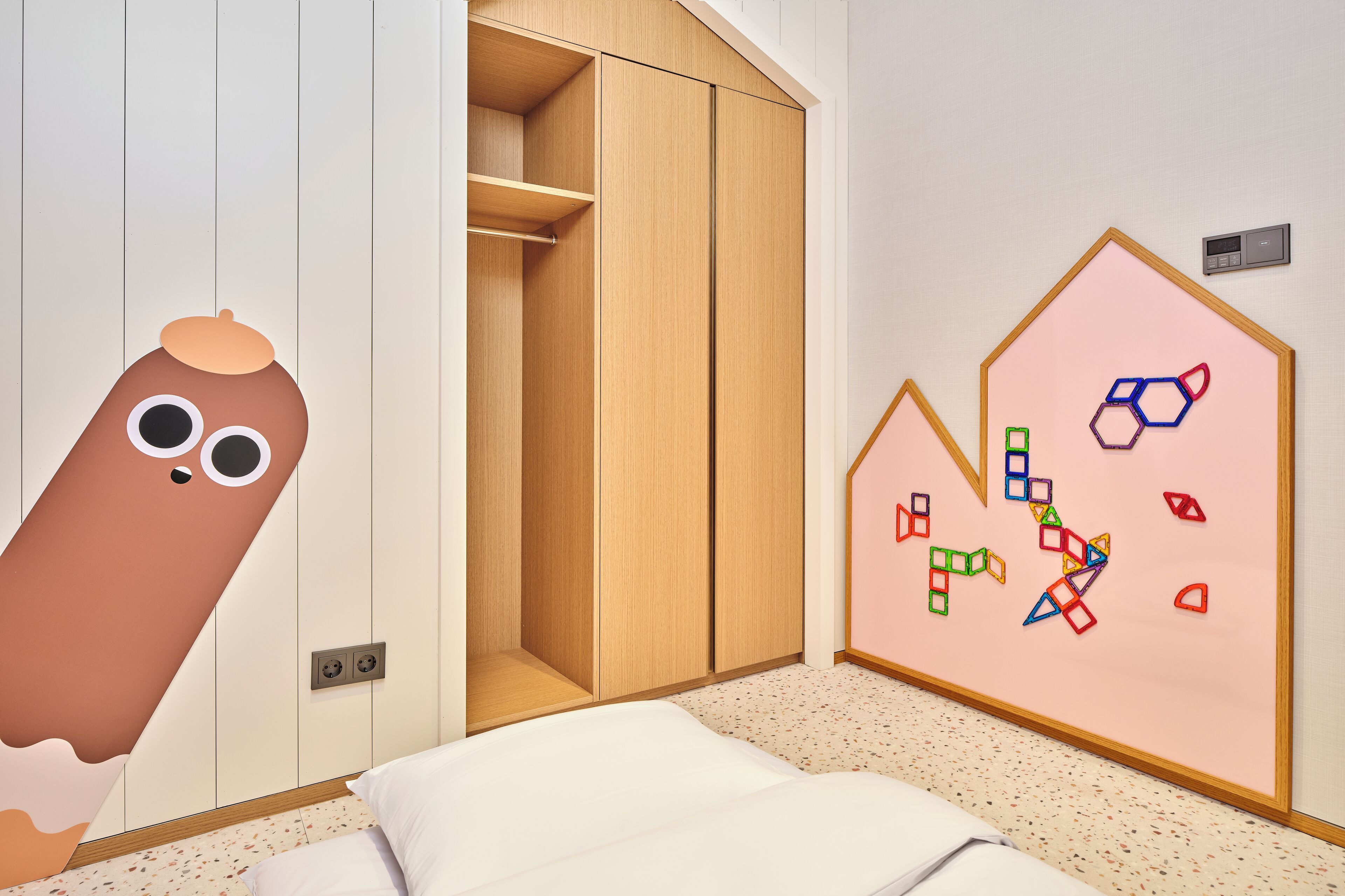 A bedroom with a wooden wardrobe, a bed with white bedding, and a wall with colorful geometric shapes.