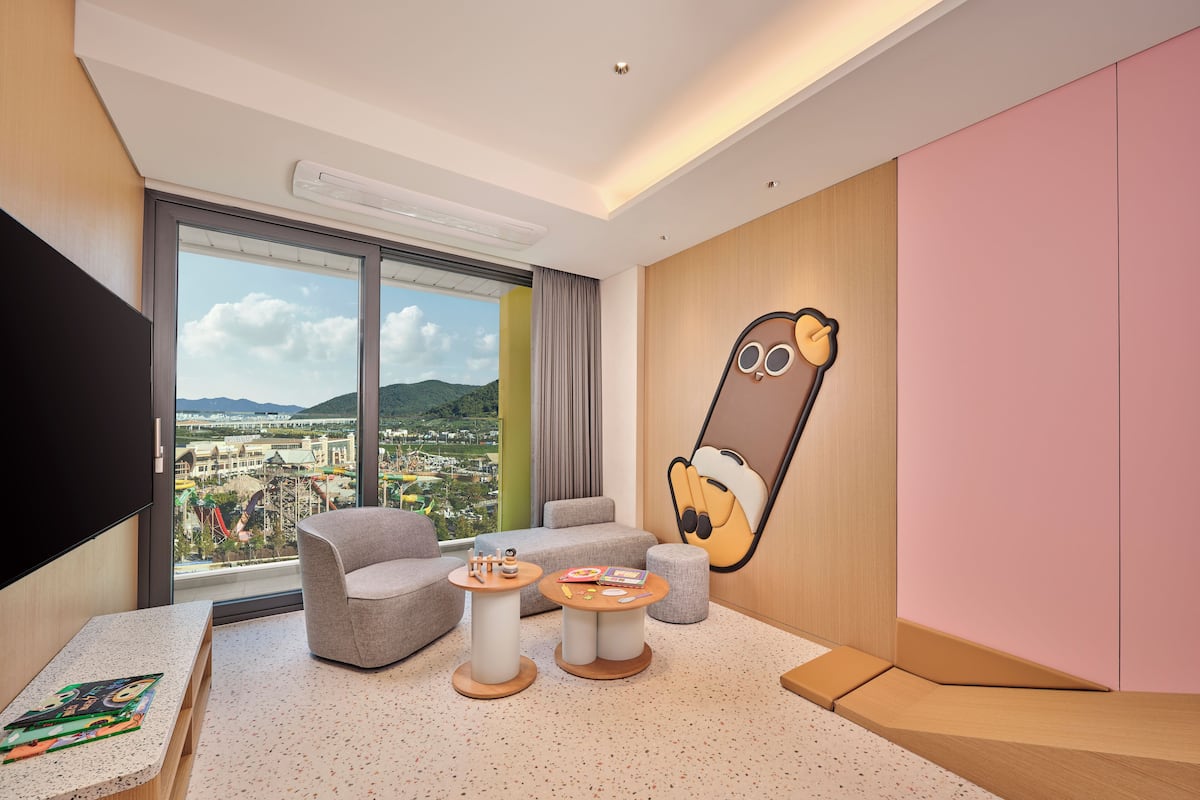 A modern hotel room with a large window offering a city view, a flat-screen TV, a comfortable seating area, and a playful wall decoration.