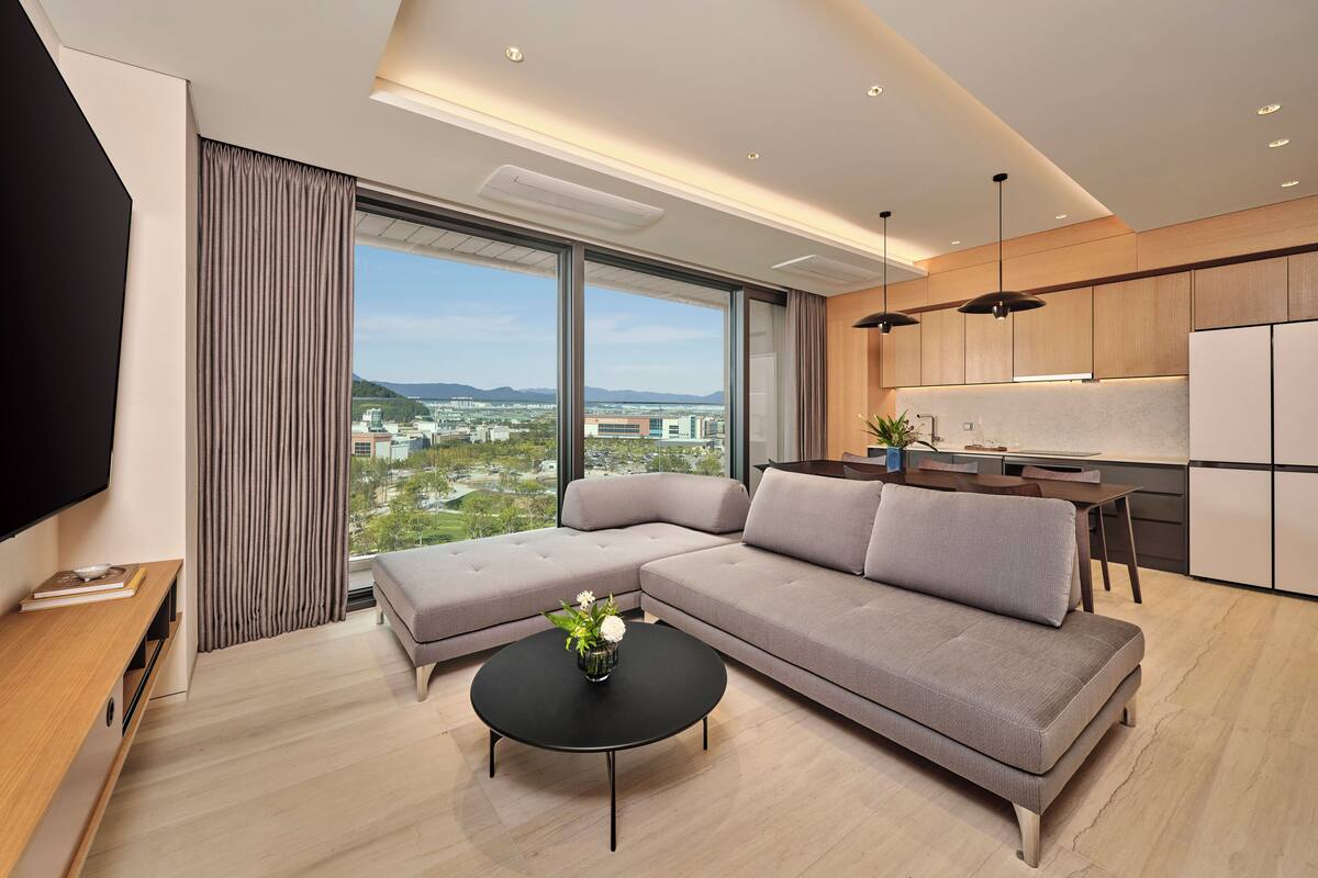 A modern living room with a sectional sofa, a black coffee table with a vase, a flat-screen TV, and a large window offering a city view.
